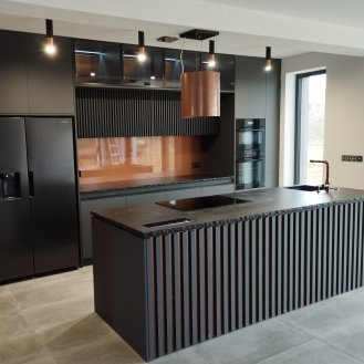 Kuchyňská linka na míru se žebrovanými dvířky v dekoru Šedá Basalt OM s ostrůvkem a kamennou pracovní deskou se štípanými hranami. Stěna je obložená laminem v dekoru Dub Sherman koňakově hnědý do které jsou vyřezaná dvířka. Většina doplňků jsou použita v měděné barvě.