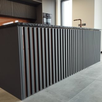 Kuchyňská linka na míru se žebrovanými dvířky v dekoru Šedá Basalt OM s ostrůvkem a kamennou pracovní deskou se štípanými hranami. Stěna je obložená laminem v dekoru Dub Sherman koňakově hnědý do které jsou vyřezaná dvířka. Většina doplňků jsou použita v měděné barvě.
